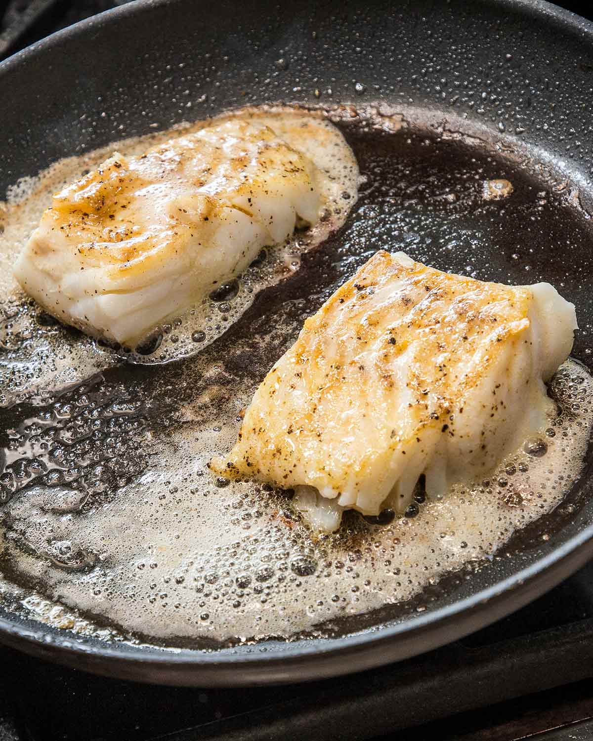 Two pieces of butter basted fish with garlic and thyme in a skillet with butter foaming around them.