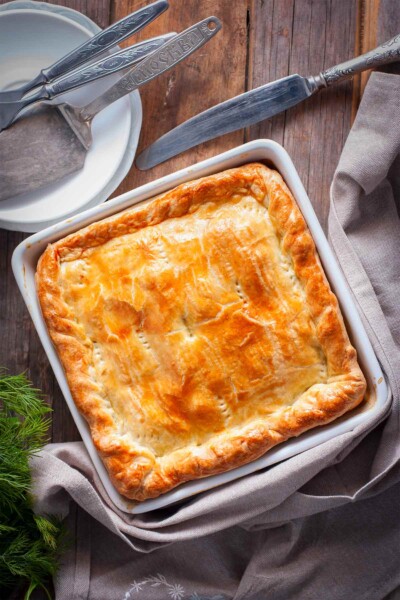 A chicken and leek pot pie in a white square baking dish with a server and forks and knife nearby.