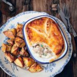 A chicken mushroom pot pie in a blue and white rectangular pie dish on a floral plate with fried potato chunks next to it.