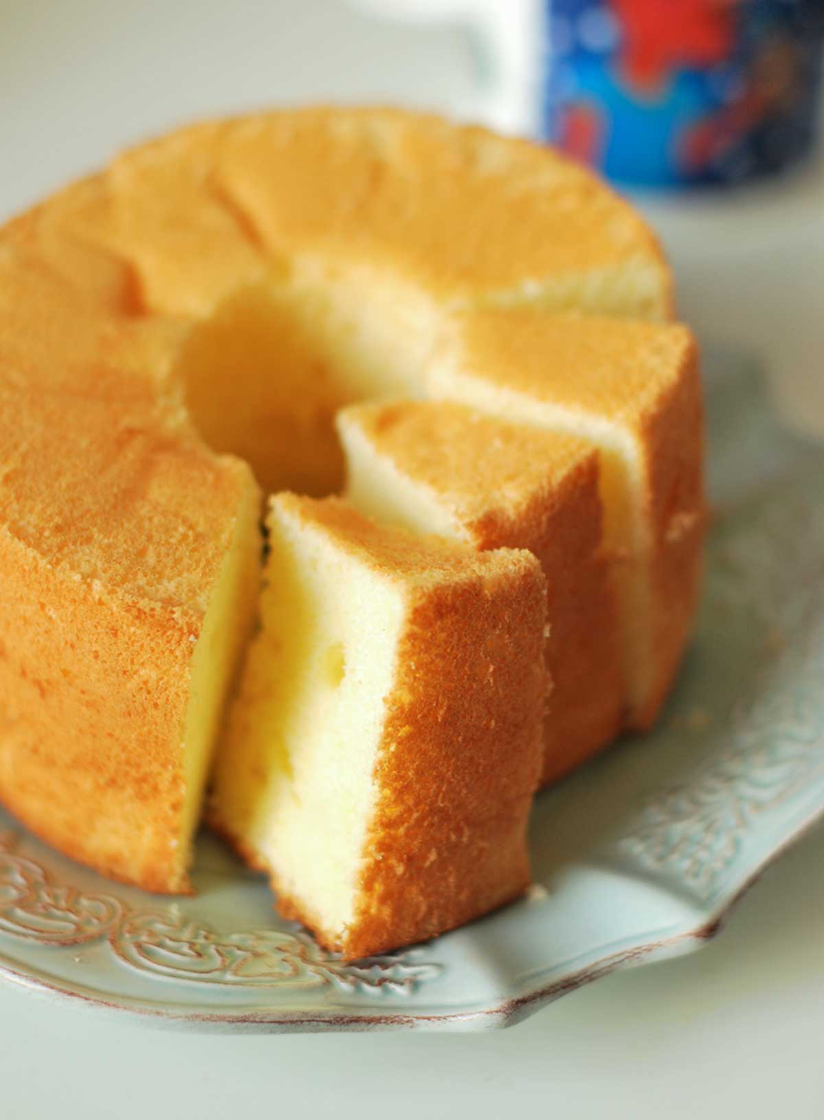 A chiffon cake with lemon icing on a white decorative plate with a few slices cut from it.