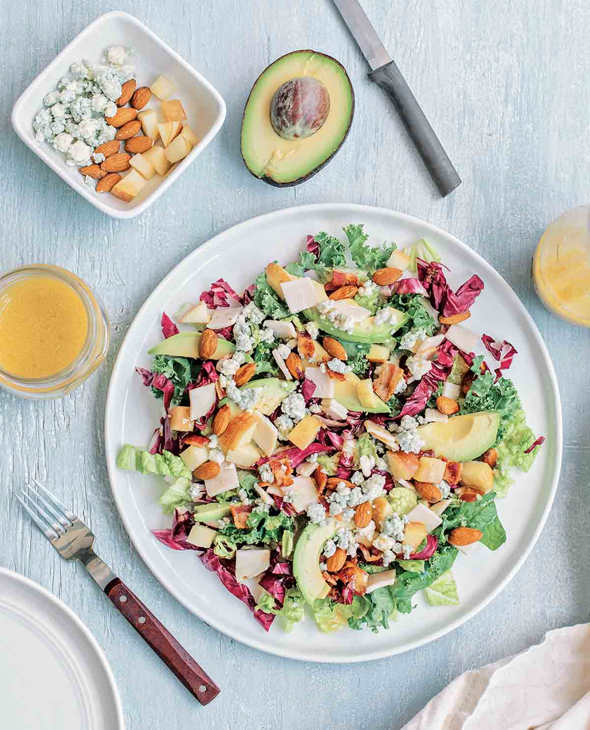 Chopped salad on a white plate, with a fork and knife, halved avocado, and dish of blue cheese, almonds, and apple on the side.