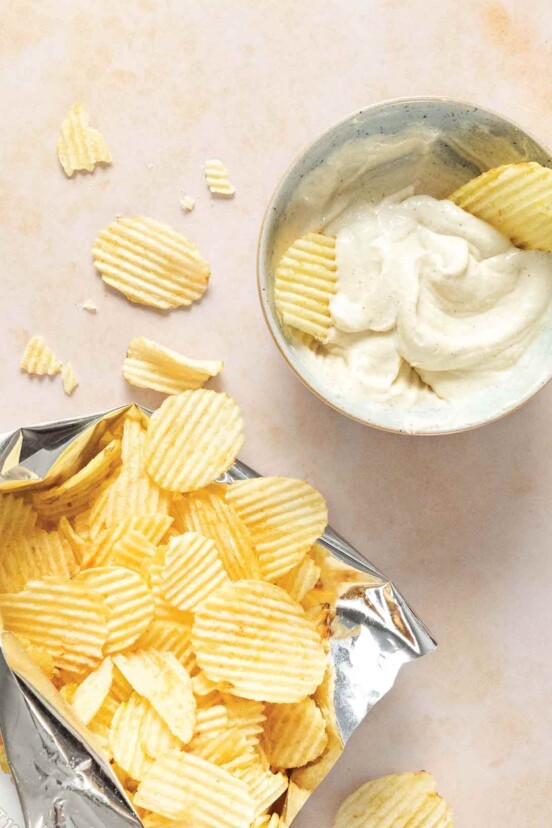 An open bag of ruffle chips next to a bowl of classic onion dip with two chips stuck in the dip.