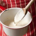 A person holding a pot of creamy stone ground cheese grits with a wooden spoon in it.