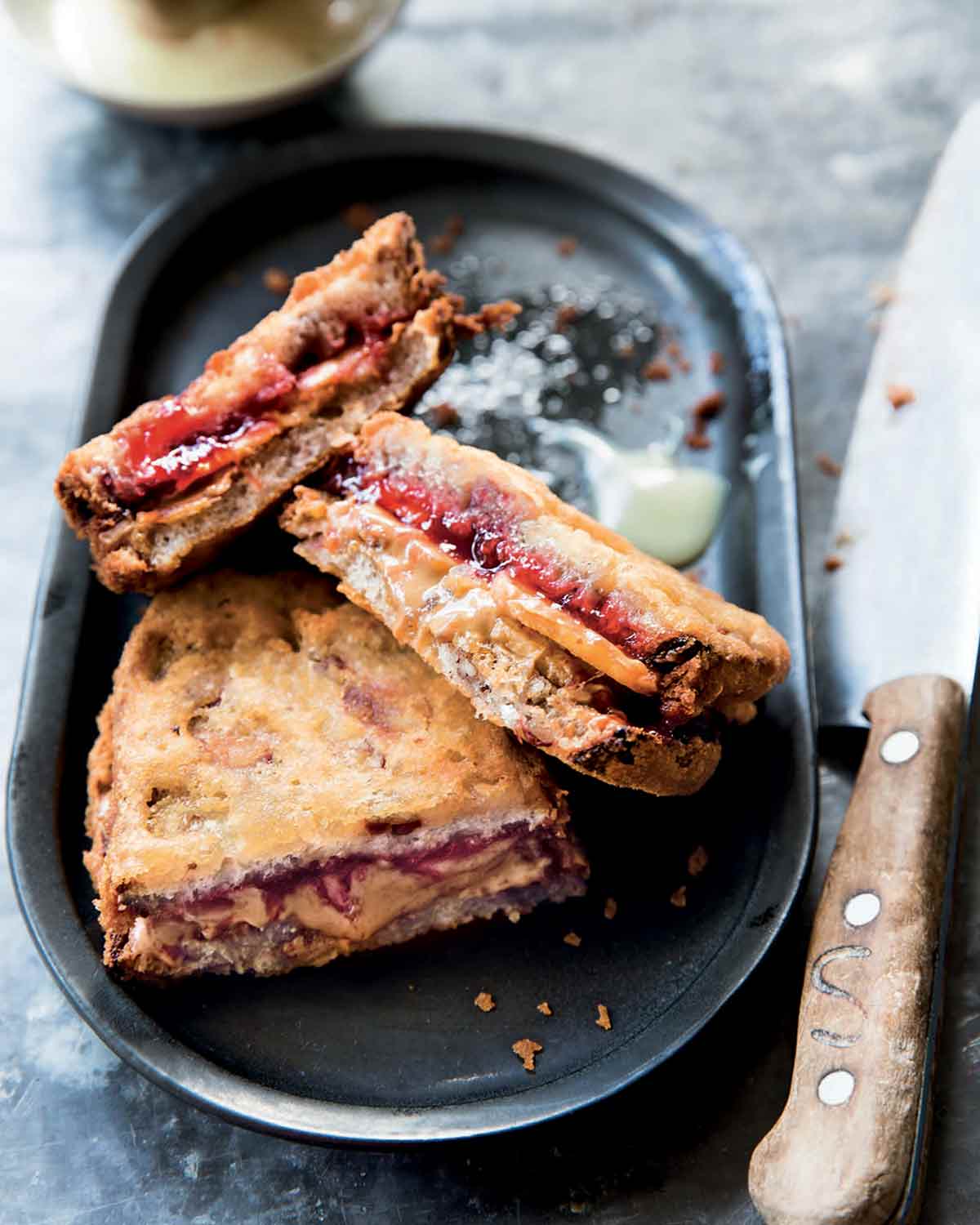 Three halves of deep-fried peanut butter and jelly sandwiches on a black oval platter with a knife on the side.