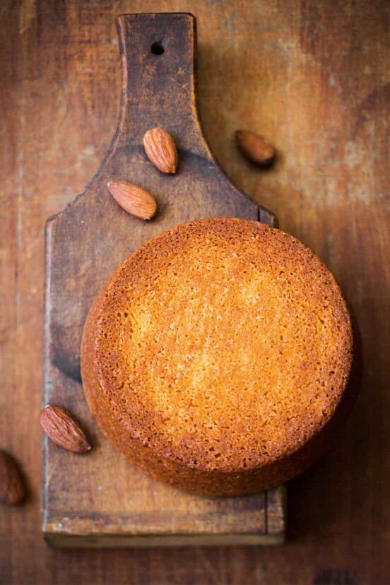 A round flourless almond cake on a wooden baking paddle with almonds scattered around it
