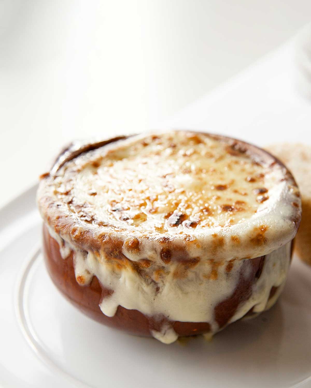 A ceramic soup bowl filled with French onion soup, and topped with melted cheese which is dripping down the side of the bowl.