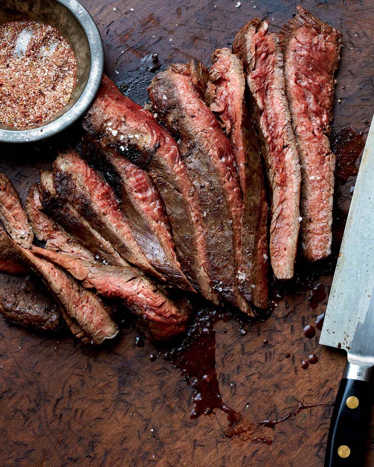 A sliced grilled flank steak with a bowl of seasoning mix and a knife on the side.