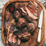 A carved grilled leg of lamb on a wooden cutting board with a knife resting beside it.