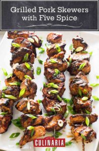 Four grilled pork skewers with five spice on a white plate, garnished with scallions.