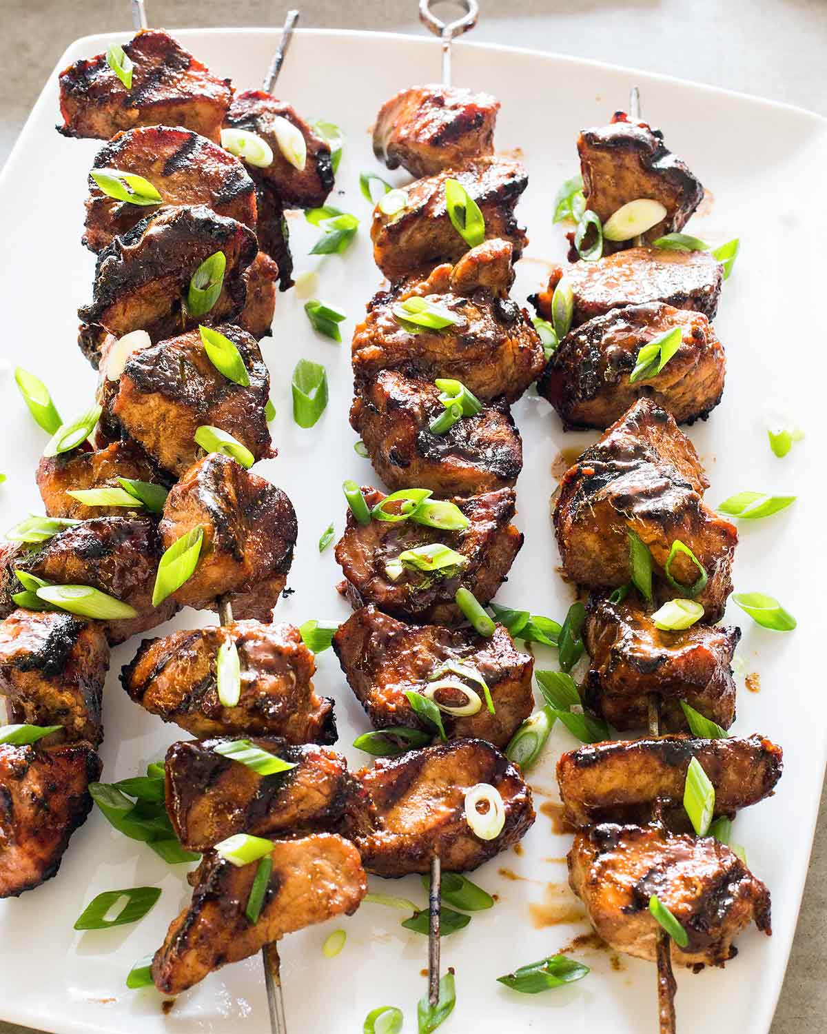 Four grilled pork skewers with five spice on a white plate, garnished with scallions.