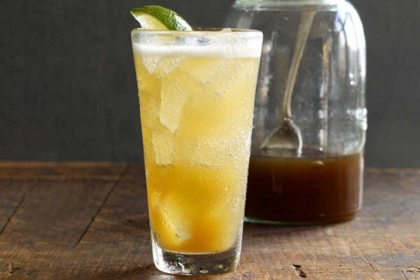 A glass of homemade ginger ale with ice and a lime wedge, and a gar of ginger syrup in the background.