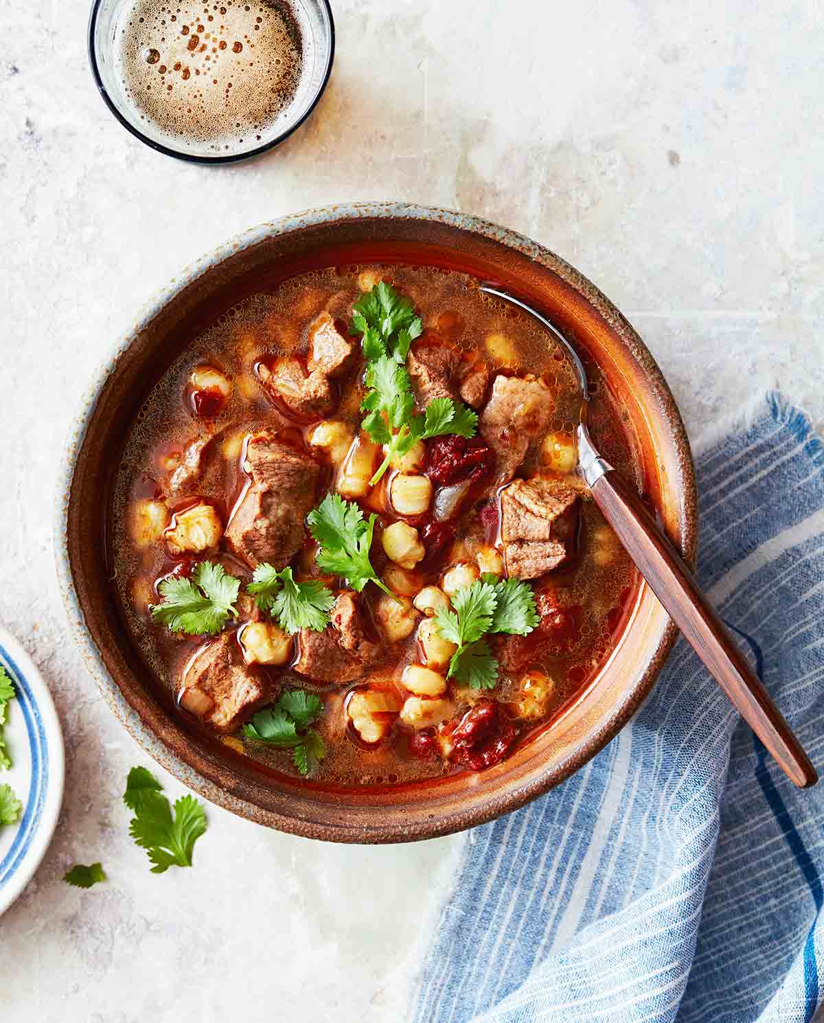 A brown bowl of Instant Pot pork stew with hominy with a spoon resting inside and a dish of cilantro and a blue napkin on the side.