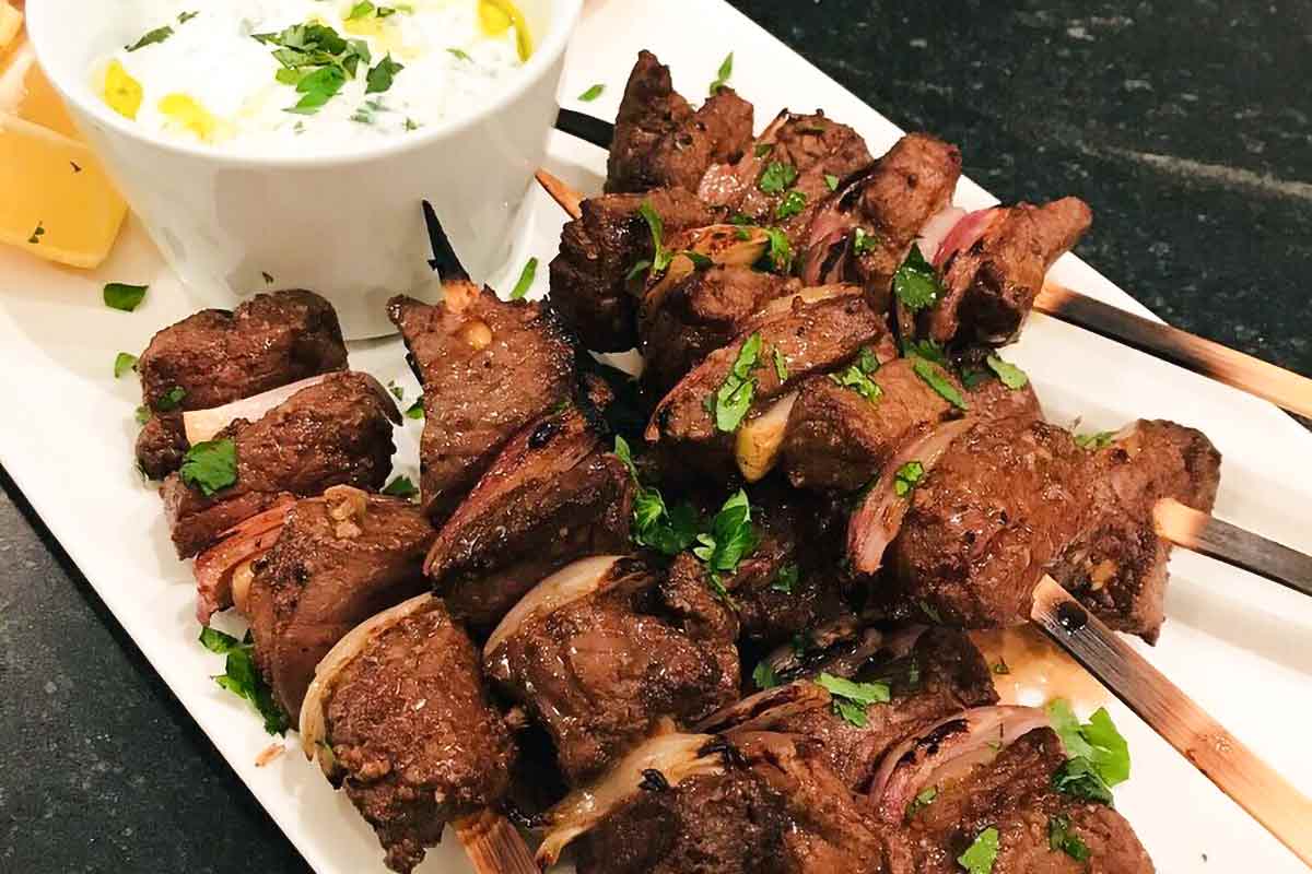 Sheet pan beef skewers piled on a white rectangular serving platter with a white dip and lemon wedges.