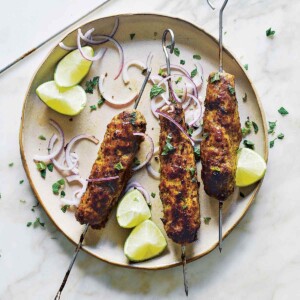 Three lamb sheekh kebab skewers on a plate with sliced red onion, lime wedges, and chopped mint leaves.
