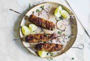 Three lamb sheekh kebab skewers on a plate with sliced red onion, lime wedges, and chopped mint leaves.