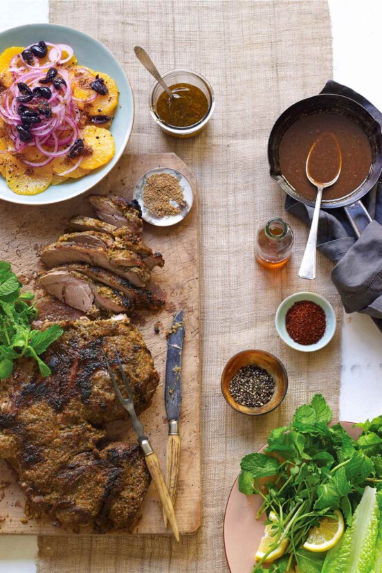 A wooden board with a partially carved leg of lamb with Moroccan spices, with side dishes and bowls of spices and gravy alongside.