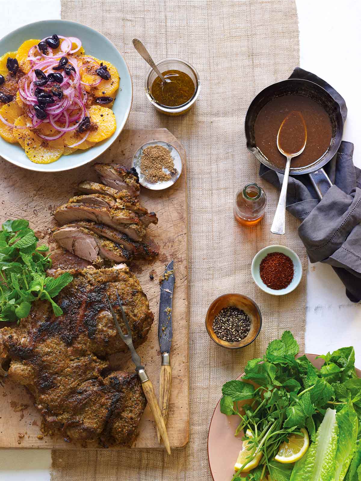 A wooden board with a partially carved leg of lamb with Moroccan spices, with side dishes and bowls of spices and gravy alongside.