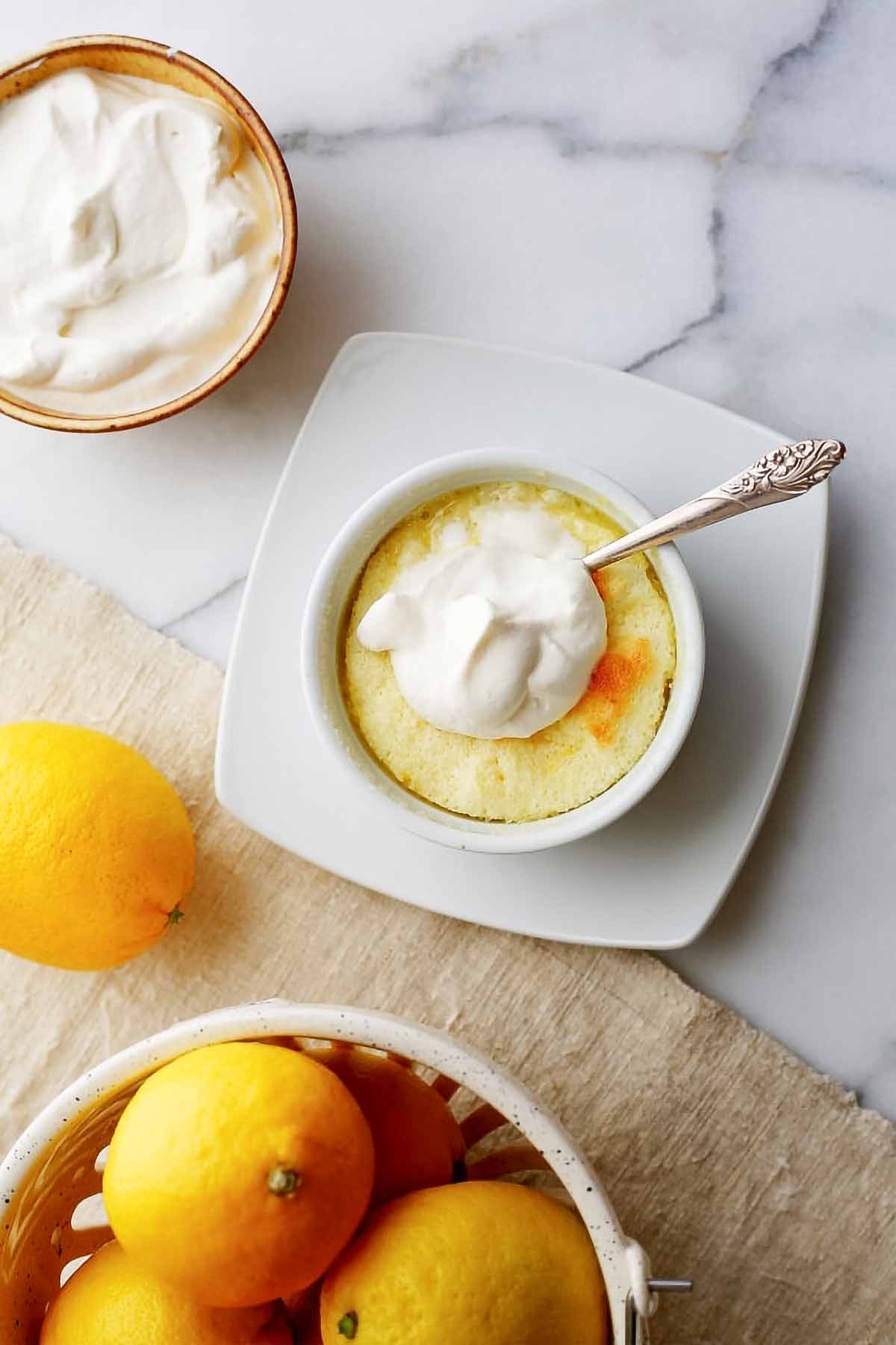 A lemon buttermilk pudding cake in a white bowl on a white plate, topped with whipped cream with a bowl of cream and basket of lemons on the side.