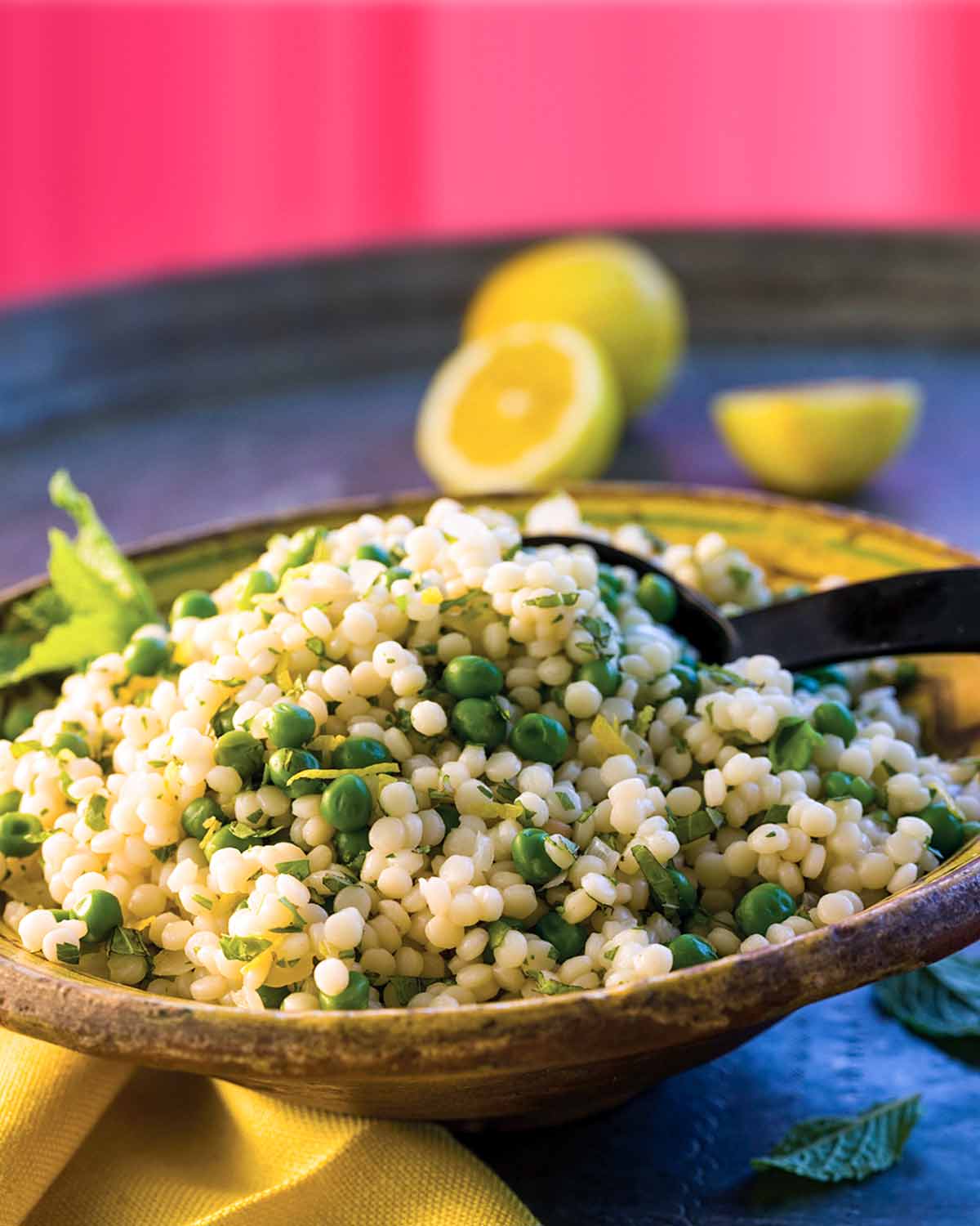 A wooden bowl filled with lemon Israeli couscous with a serving spoon inside and some lemon halves in the background.