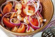 A brown bowl filled with marinated shrimp, tomato, and onion sallet, on a white and purple towel with a slotted spoon and half of a red onion resting on a plate beside the salad.