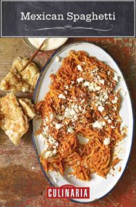 A platter of Mexican spaghetti, topped with crumbled cheese with a torn piece of bread and white sauce on the side.
