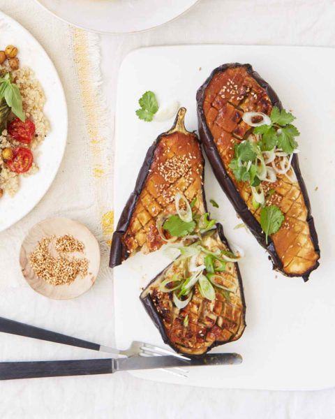 Two halves of miso-glazed eggplant sprinkled with cilantro and scallions with a fork and knife and bowl of sesame seeds beside them.