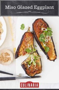 Two halves of miso-glazed eggplant sprinkled with cilantro and scallions with a fork and knife and bowl of sesame seeds beside them.