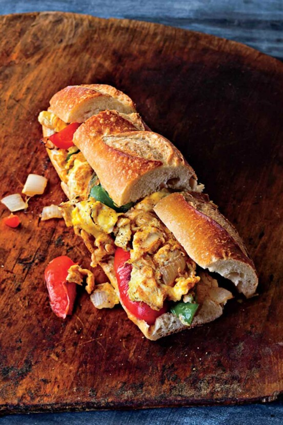 A pepper and egg sandwich on a wooden table with pieces of pepper and onion falling out.