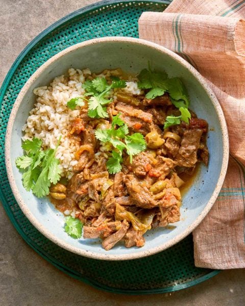 A bowl of Puerto Rican-style ropa vieja with rice and cilantro on a green mesh serving tray with a linen napkin beside it.