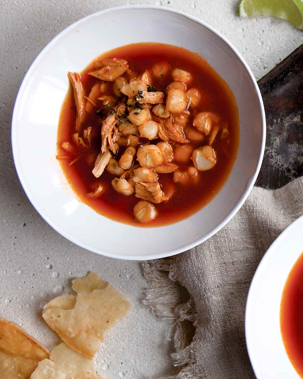 Two white bowls filled with red posole with pieces of tortilla and lime wedges on the side.