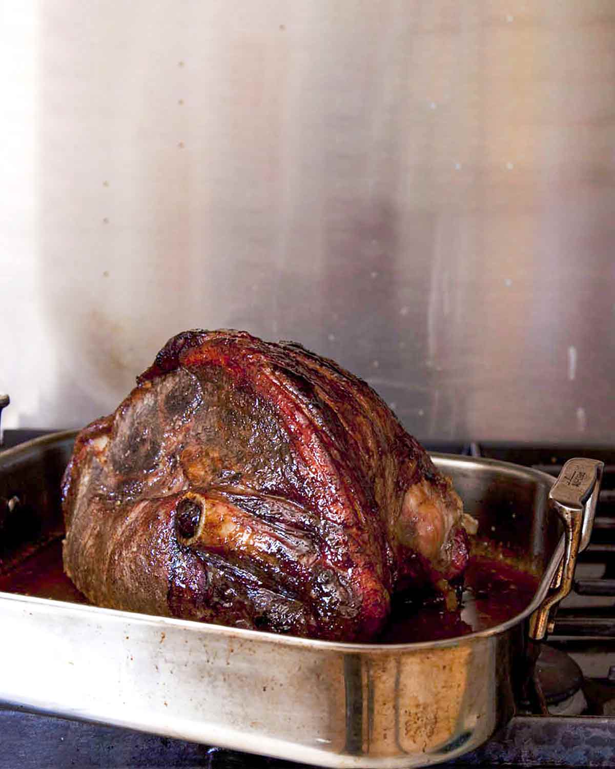 A whole roasted fresh ham with maple spice glaze in a roasting pan on a stovetop burner.