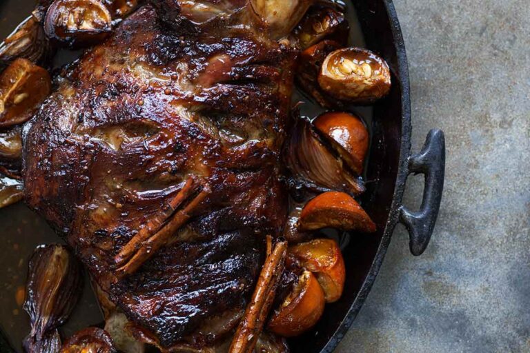 A slow roasted lamb shoulder in an oval cast iron dish with orange wedges.