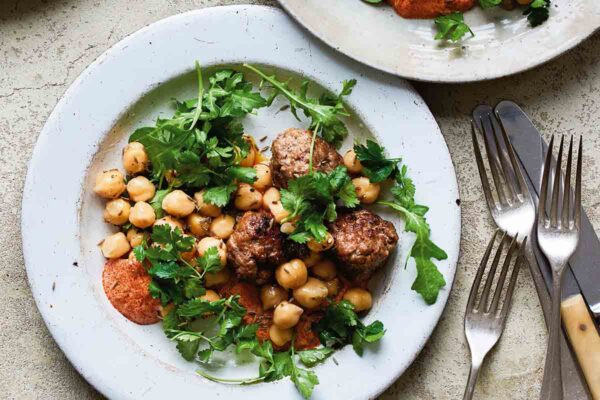 Three plates topped with spicy lamb meatballs, chickpeas, and arugula with three forks and two knives on the side.
