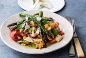 A white plate topped with a spring panzanella made with tomatoes, bread, asparagus, and Parmesan. A small plate of additional shaved Parmesan sits beside it.