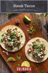 Two steak tacos on a wooden board with lime wedges, onion, and cilantro scattered around.