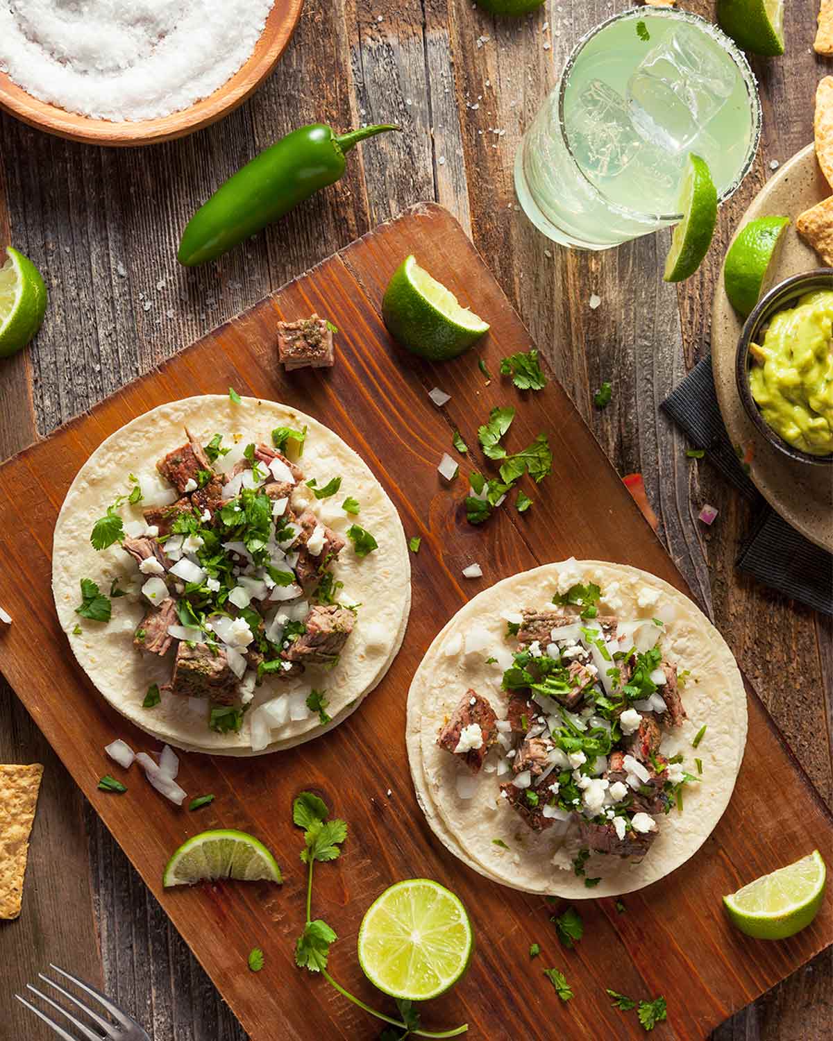 Two steak tacos on a wooden board with lime wedges, onion, and cilantro scattered around.