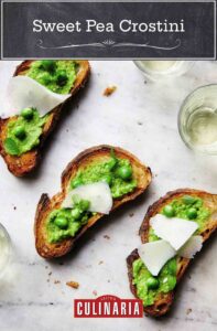Three sweet pea crostini topped with pecorino shavings on a marble surface with glasses of white wine nearby.