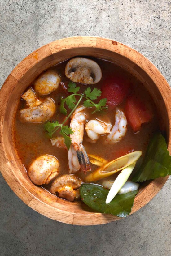 A wooden bowl filled with tom yum goong soup with mushrooms, lemongrass, shrimp, and cilantro in a broth.