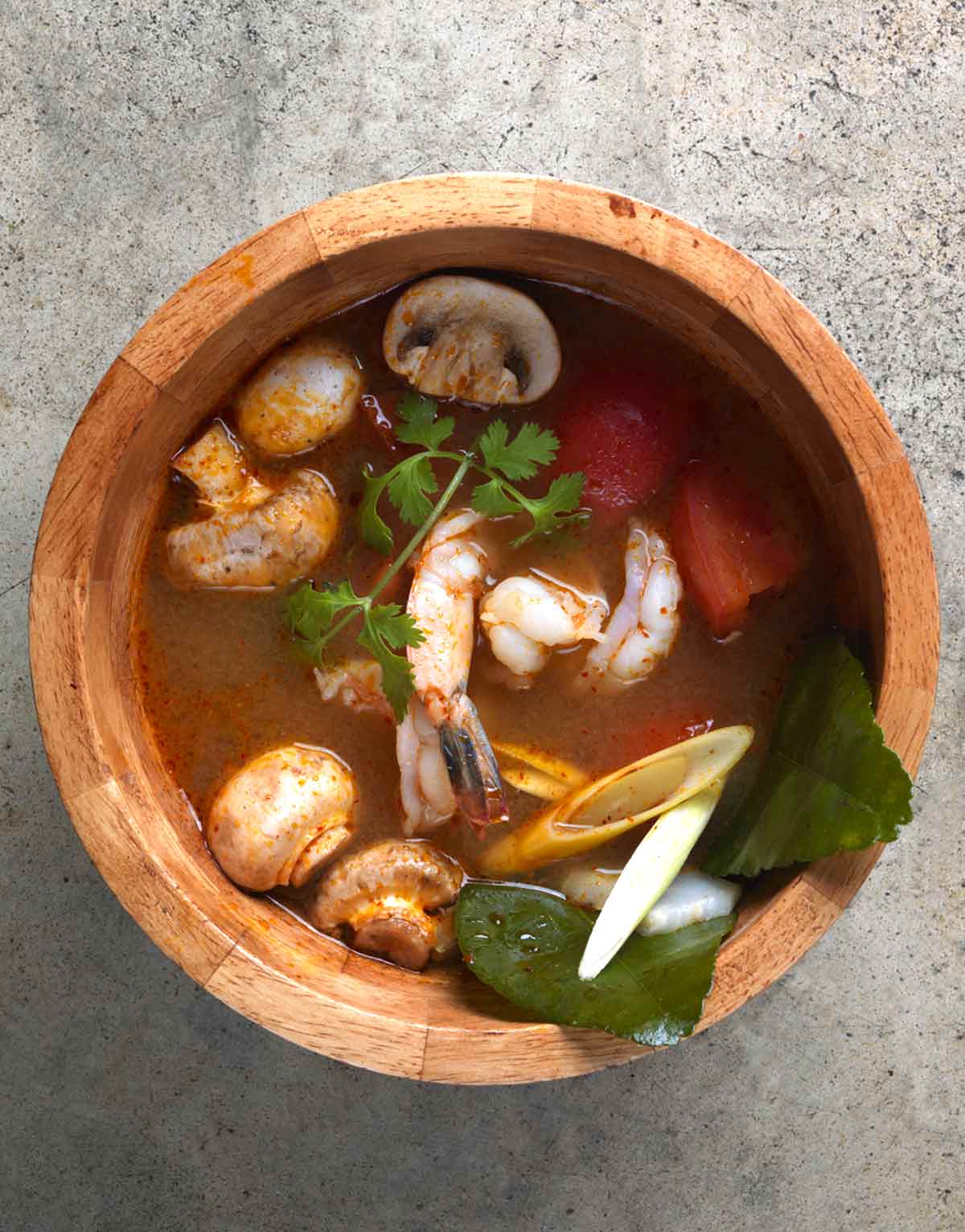 A wooden bowl filled with tom yum goong soup with mushrooms, lemongrass, shrimp, and cilantro in a broth.