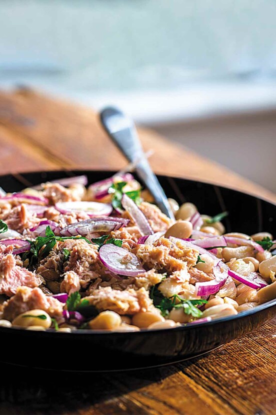 Tuna salad with white beans, red onion, and parsley in a black bowl with a fork, on a wooden table.