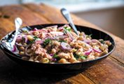 Tuna salad with white beans, red onion, and parsley in a black bowl with a fork and spoon, on a wooden table.