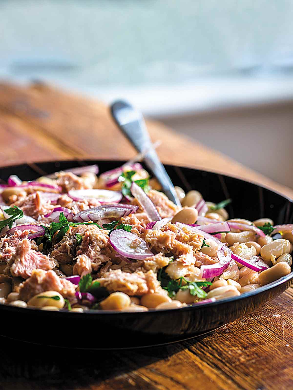Tuna salad with white beans, red onion, and parsley in a black bowl with a fork, on a wooden table.