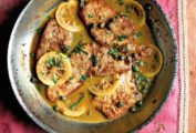 Four veal cutlets in a white wine lemon sauce with lemon slices and capers in a skillet for veal piccata.