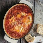 An oval casserole dish of acquacotta - eggs poached in a tomato and onion sauce -- with a piece of bread, plate, and utensils on the side.