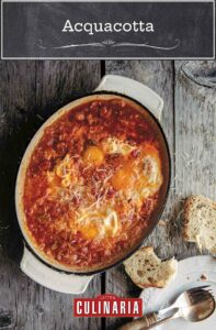 An oval casserole dish of acquacotta - eggs poached in a tomato and onion sauce -- with a piece of bread, plate, and utensils on the side.