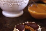 A white bowl filled with alfajores, and one halved one in front of the bowl with a bowl of dulce de leche beside