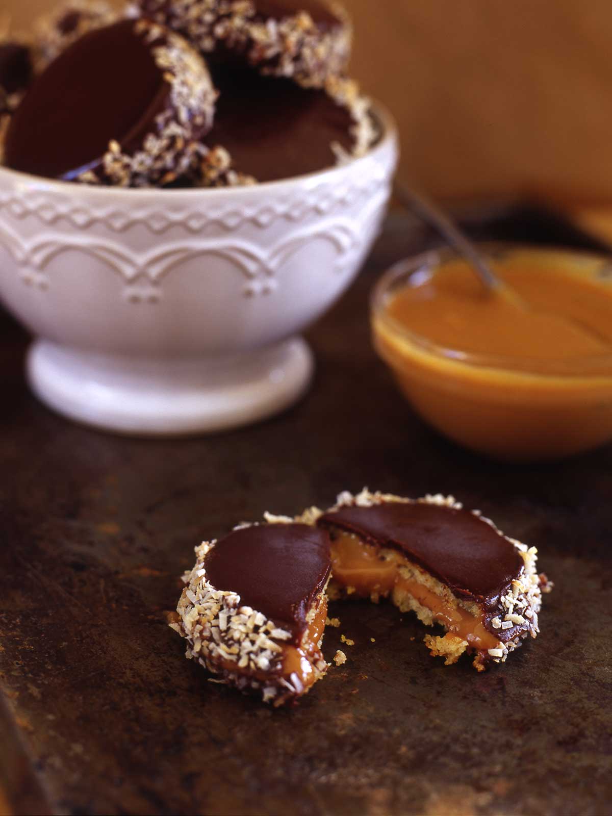 A white bowl filled with alfajores, and one halved one in front of the bowl with a bowl of dulce de leche beside