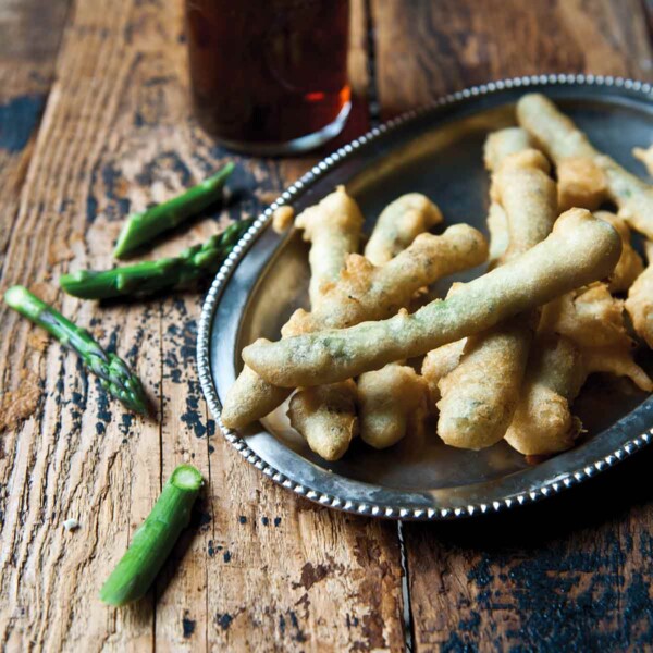 A silver platter filled with asparagus tempura with some pieces of asparagus and a glass of beer on a wooden table.