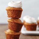 Three banana bread muffins stacked on top of each other with chai whipped cream on top and more muffins on a plate in the background.