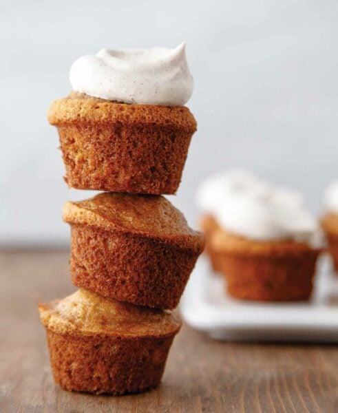 Three banana bread muffins stacked on top of each other with chai whipped cream on top and more muffins on a plate in the background.